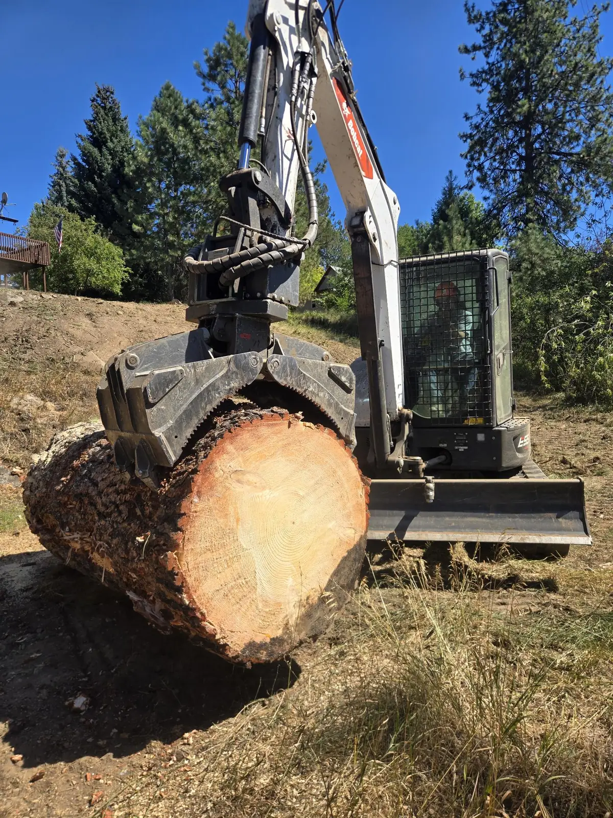 Excavator grapple lifting a large fresh-cut log on a cleared hillside lot